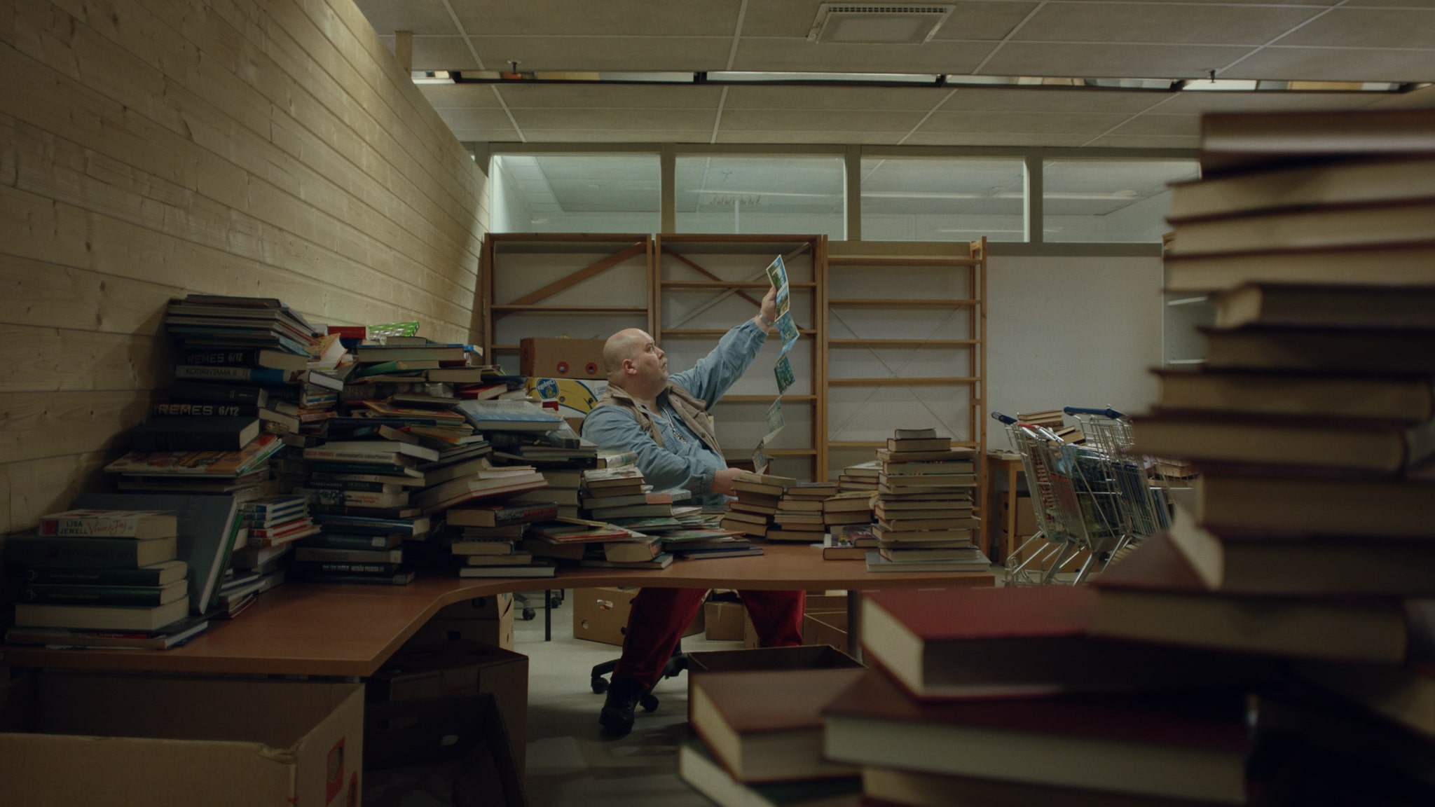 En man sitter i ett dovt belyst rum omringad av stökigt travade böcker.