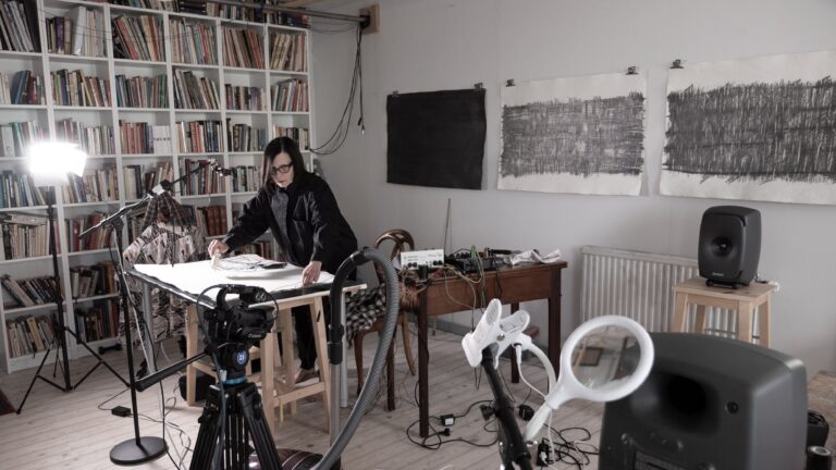 Ann Rosén i ett ljust rum fyllt av stativ, kablar och övrig elektronik medan hon framför Drawing Session. På väggen bakom henne hänger stora teckningar.