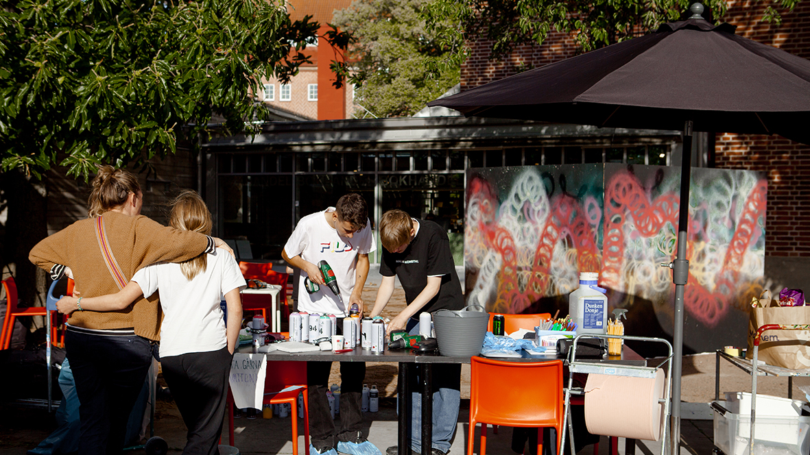 Utomhusverkstad en sommardag. Ungdomar står runt ett uppdukat bord av olika verktyg och sprayfärg och skapar.