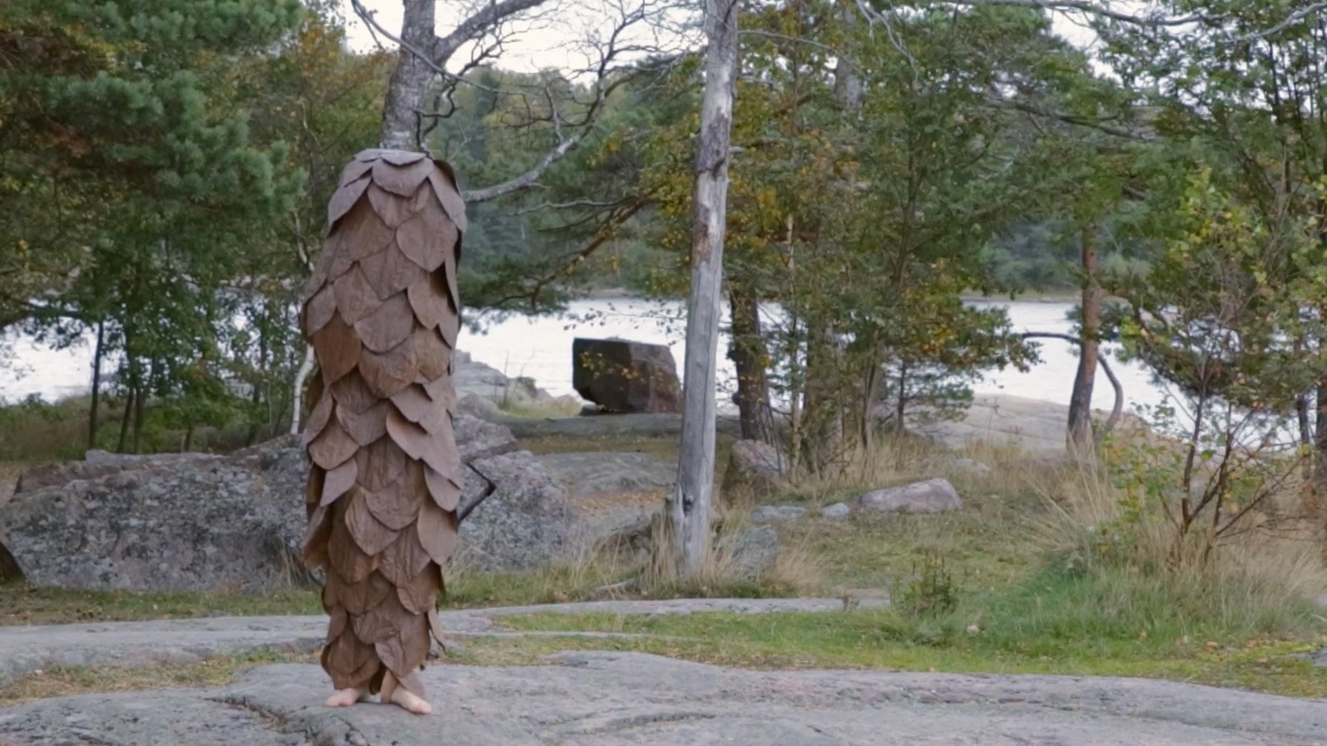 Grankotte i mänsklig storlek står på en klippa. Det är konstnären Ingela Ihrman som är utklädd till en kotte. Hennes fötter sticker ut längst ner på kostymen.