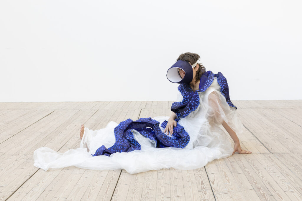 Ingela Ihrman sittandes på golvet i ett av hennes verk som ser ut som en stor mussla. Dräkten är av ljus tyll och ett mörkblått tyg som slingrar sig runt och uppepå dräkten. Hon har en knuten skärm för ansiktet som hör till dräkten.