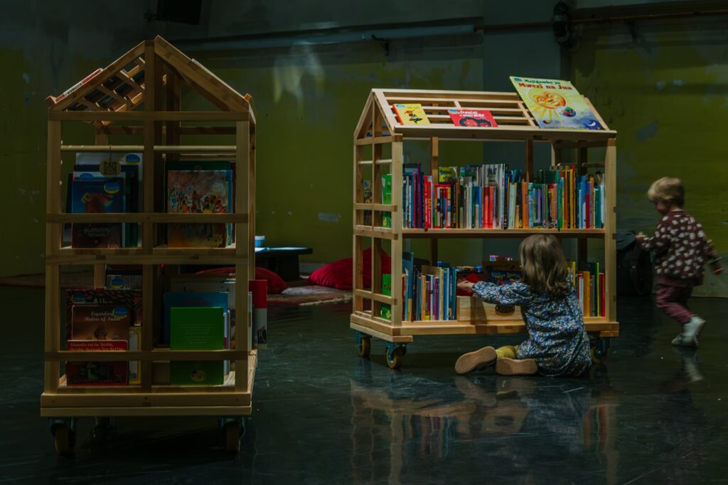 Två träkonstruktioner formade som små hus och fyllda med böcker för barn och unga. Framför dem sitter två barn och bläddrar i böcker.