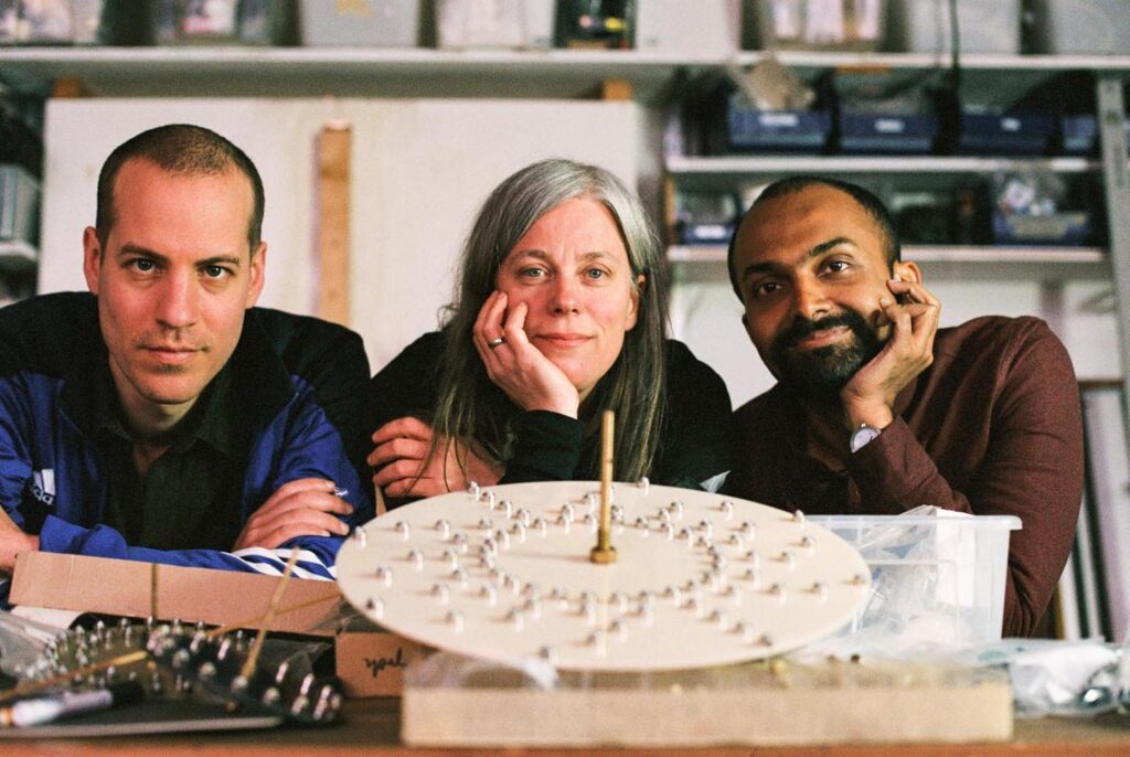 En gruppbild av tre konstnärer som sitter bredvid varandra med ett verk som föreställer en planet skapad i plywood framför sig.