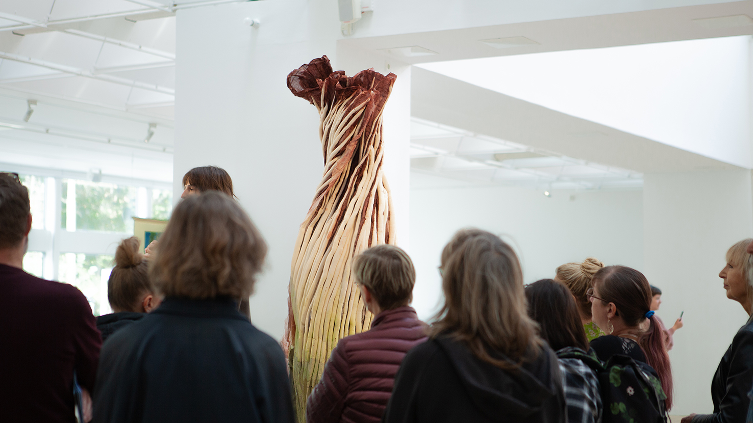 A crowd gathers around an artwork depicting a human-sized flower and a guide.