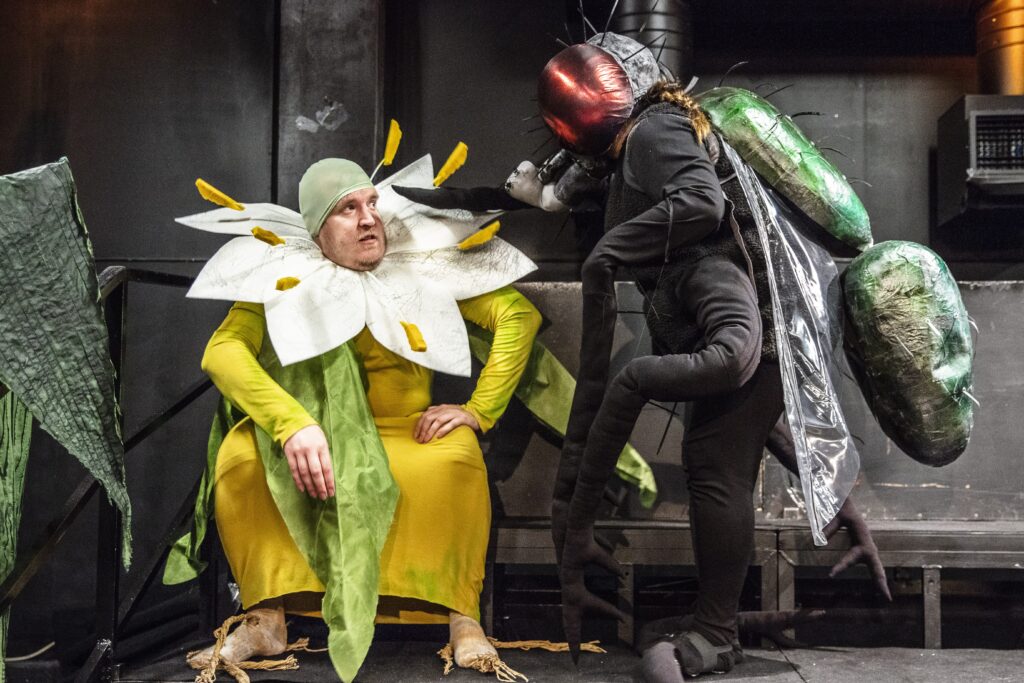 Två skådespelare utklädda till en blomma och en spyflyga sitter och samtalar. Kostymerna är del av pjäsen Vilja är ni? som skapats av konstnären Ingela Ihrman.
