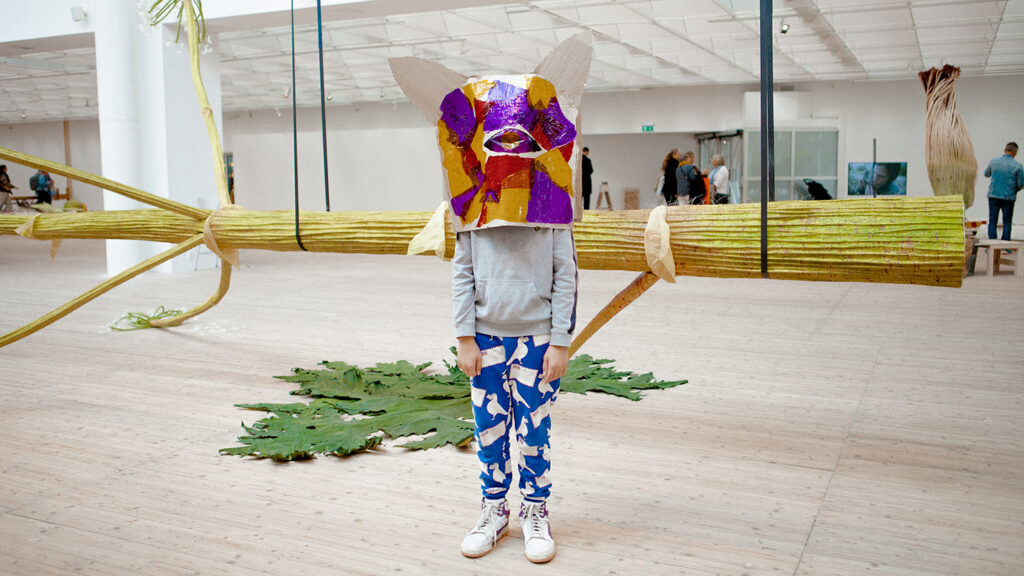 En inspirerande bild av ett barn står med en färgglad mask framför en stor skulptur av en blomsterstjälk.