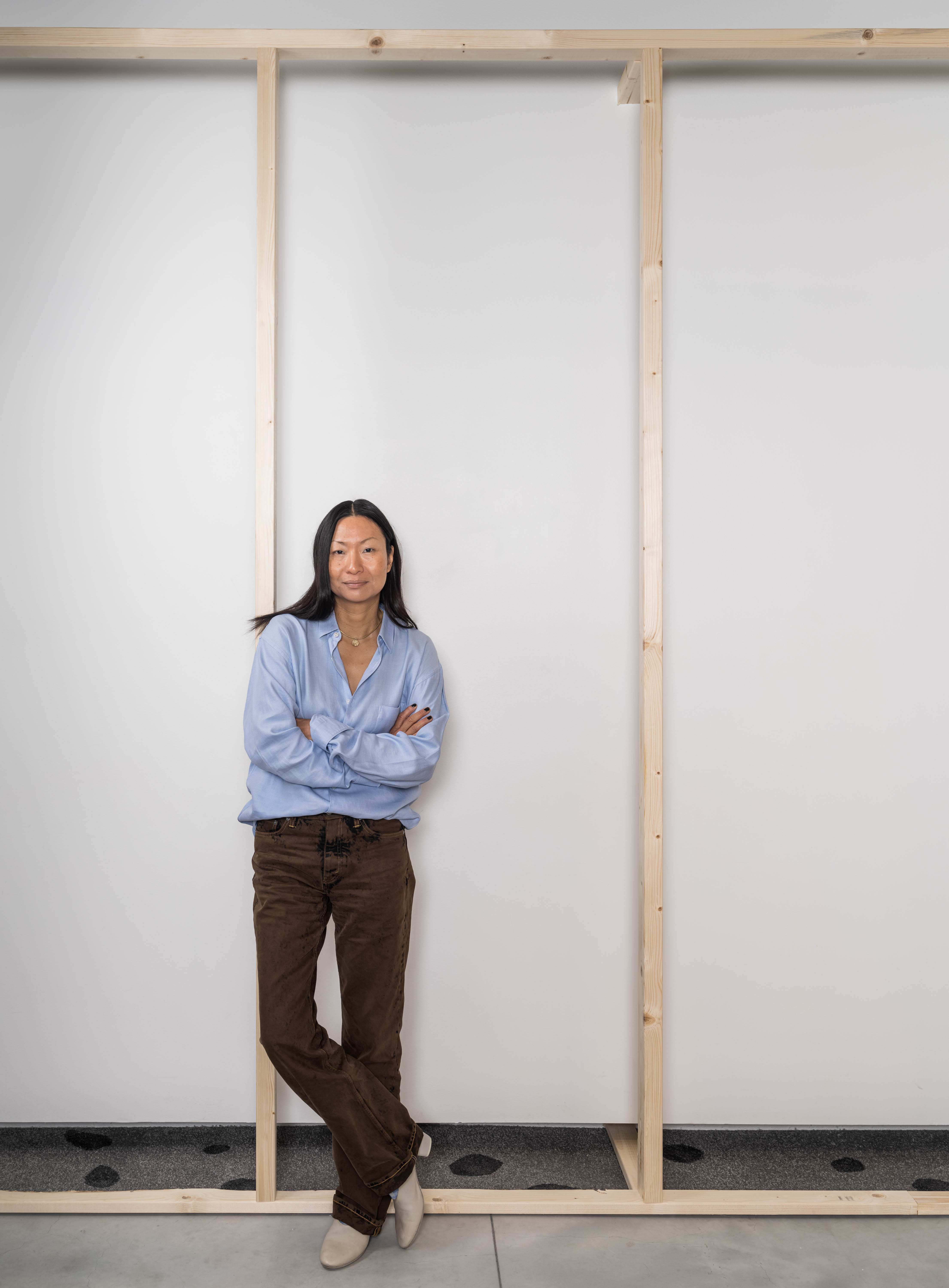 Konstnären Lisa Tan poserar med armarna i kors framför en träinstallation. Hon är ikäldd ljusblå skjorta och bruna kostymbyxor.