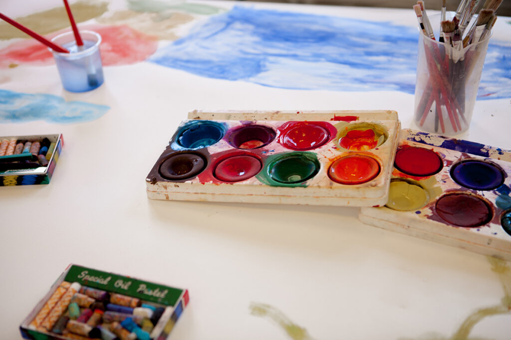 Enn inspirernade bild med ett bord med konstnärsmaterial så som vattenfärger i glada färger och kritor i askar.