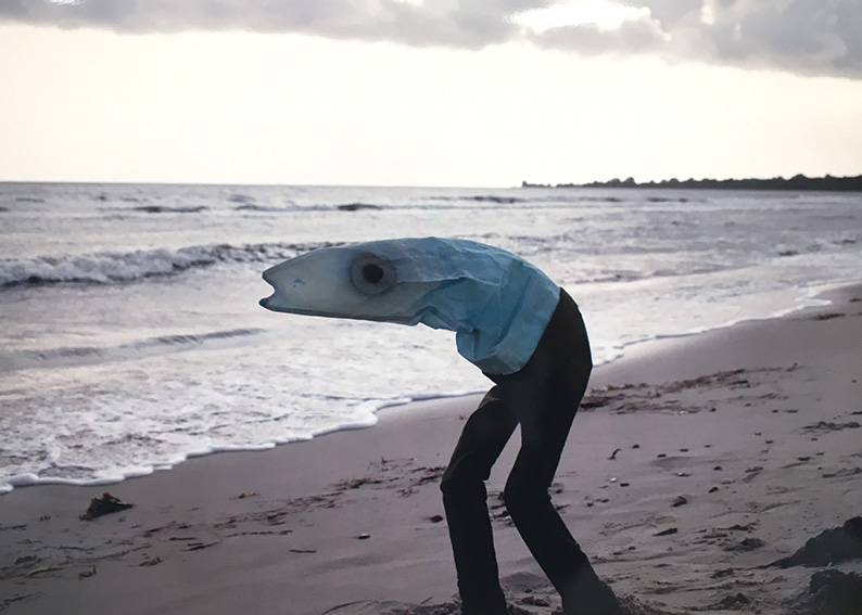 Bild av person som är utklädd till hälften fisk och hälften människa och står på en strand.