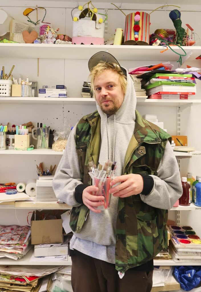 Konstnären Max står i konsthallens verkstad framför hyllor fulla av pysselmaterial och en burk pennor i handen.