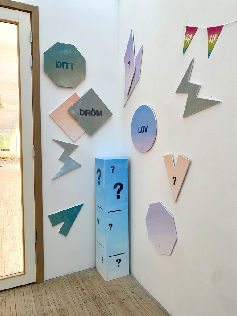 A wooden box with a painted question mark and figures mounted on the wall encourages visitors to submit requests for dream activities in the art hall's workshop during the Christmas break.