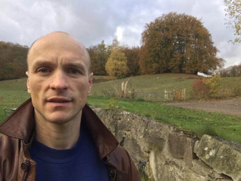 Bild av Karl Katz Lydén ute i en hage en vårdag med kullar och slätter i bakgrunden.