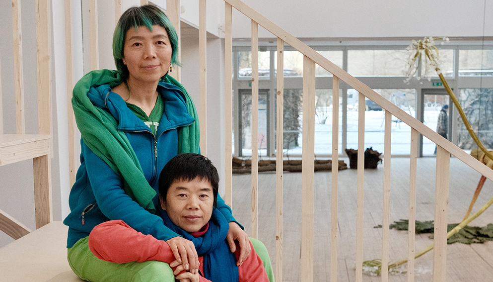 De två kinesiska konstnärerna Shi Tou och Mingming sitter tätt intill varandra och håller om varandra på Malmö Konsthall. De ser lugna och glada ut.