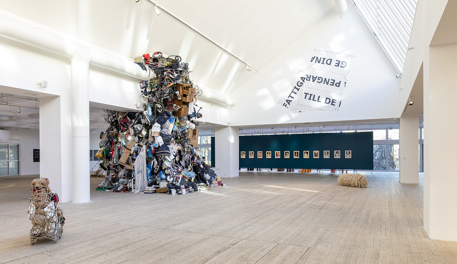 Overview of the exhibition hall. In focus, a piece resembling a large pile of debris constructed from belongings from several homes, such as beds and baby carriages. The pile is approximately five by seven meters in circumference.