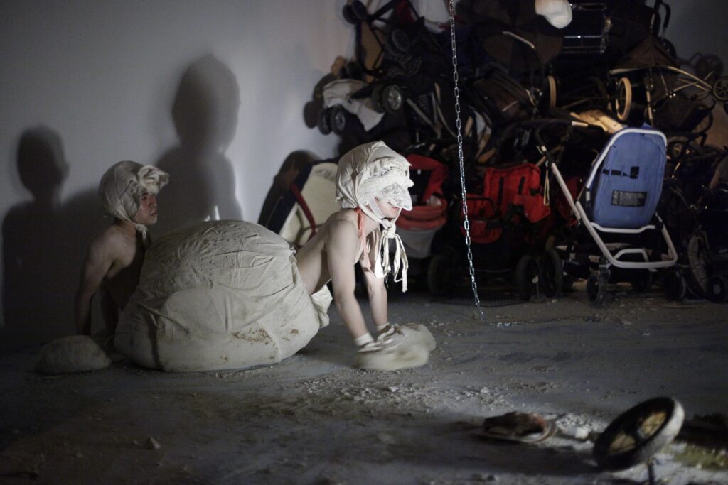 Image from a performance in a dark room with a person dressed in a cumbersome dress in front of a pile of furniture and items.