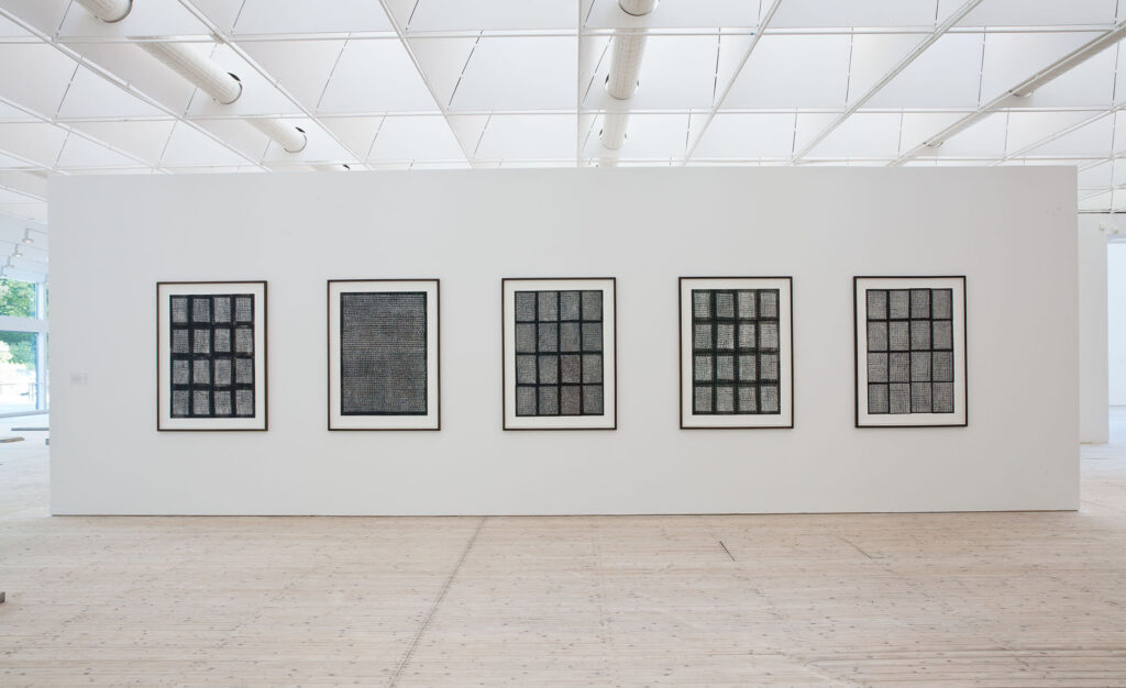 En vit vägg på konsthallen med fem stora inramade verk som ser ut som fönster med galler i nyanser av svart och grått.