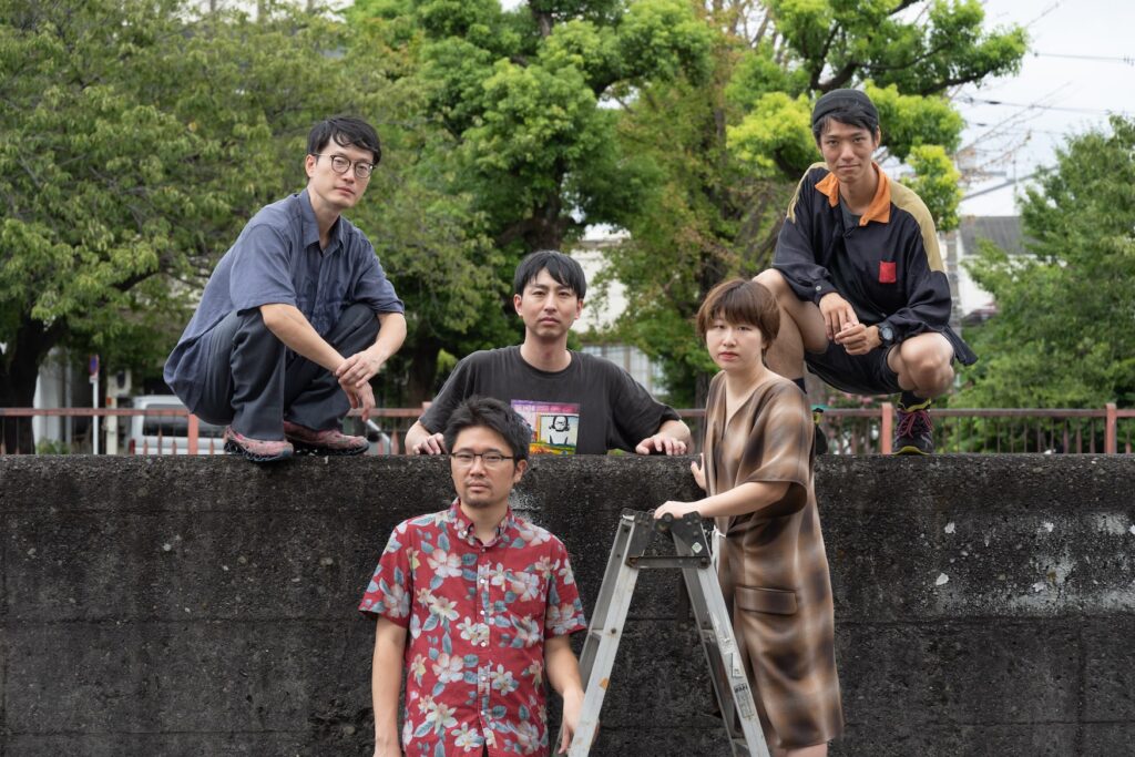 Fem japanska konstnärer i kollektivet OLTA på en gruppbild utomhus vid en mur och gröna träd i bakgrunden.