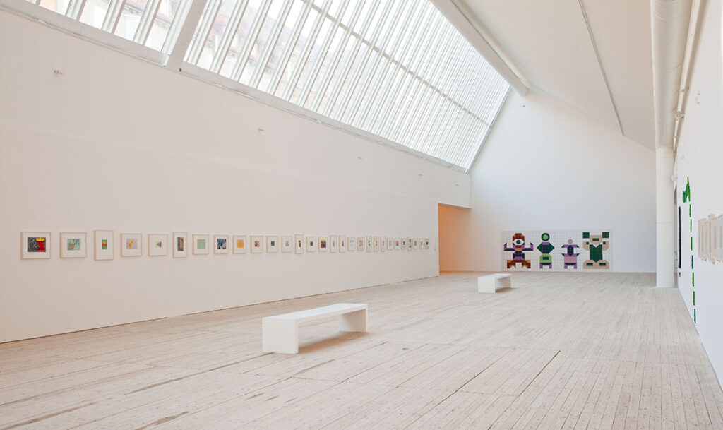 En överblick över konsthallens rum med ljusinsläpp från taket och en vägg med en lång rad små målningar och ett större verk utfört i kakel.