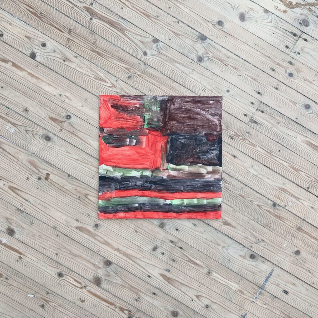 En tavla som ser ut att vara målad med vattenfärg. Röda, bruna, svarta och gröna svepande penseldrag täcker hela tavlan. Tavlan ligger på ett trägolv.
