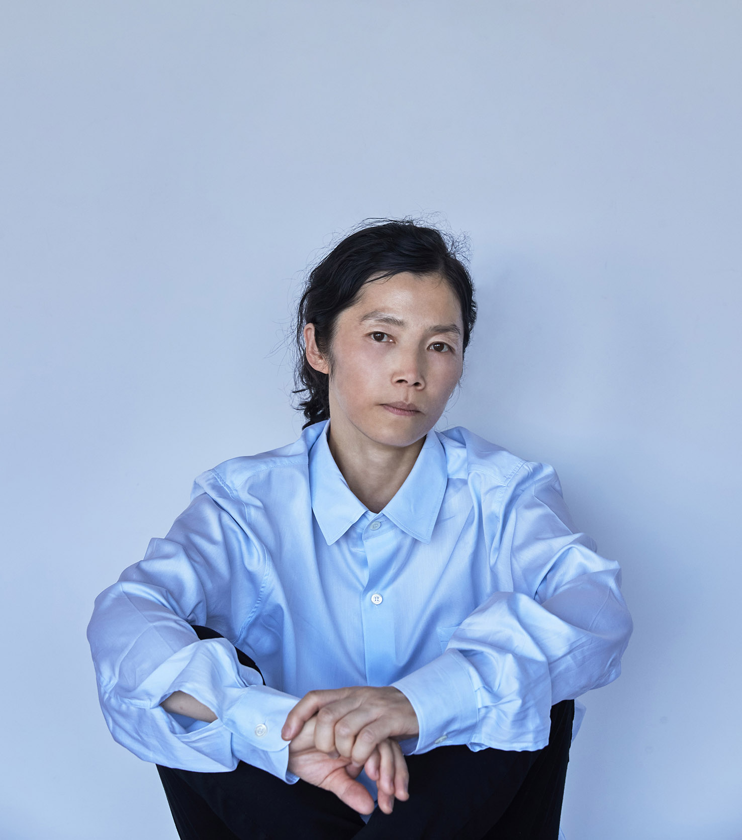Konstnären Koo Jeong A sitter framför en ljusblå vägg i en nlå skjorta.