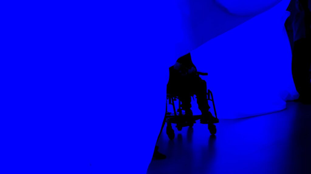 Ett rum som badar i blått ljus med en person i rullstol längst in. Rummet är omslutet av mjuka tyger formade som en grotta.