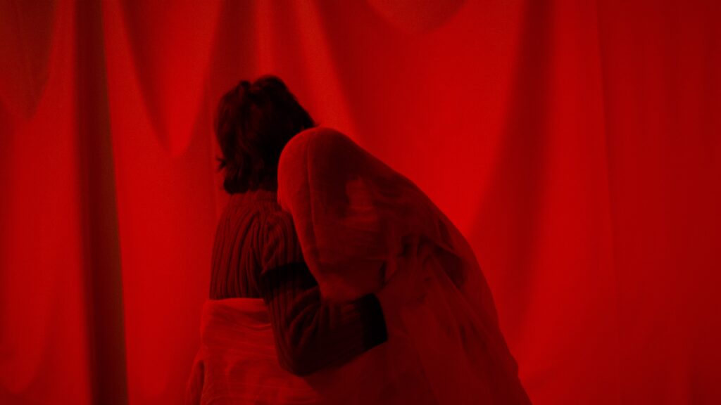 En person och en figur omfamnar varandra i ett helt rött rum.