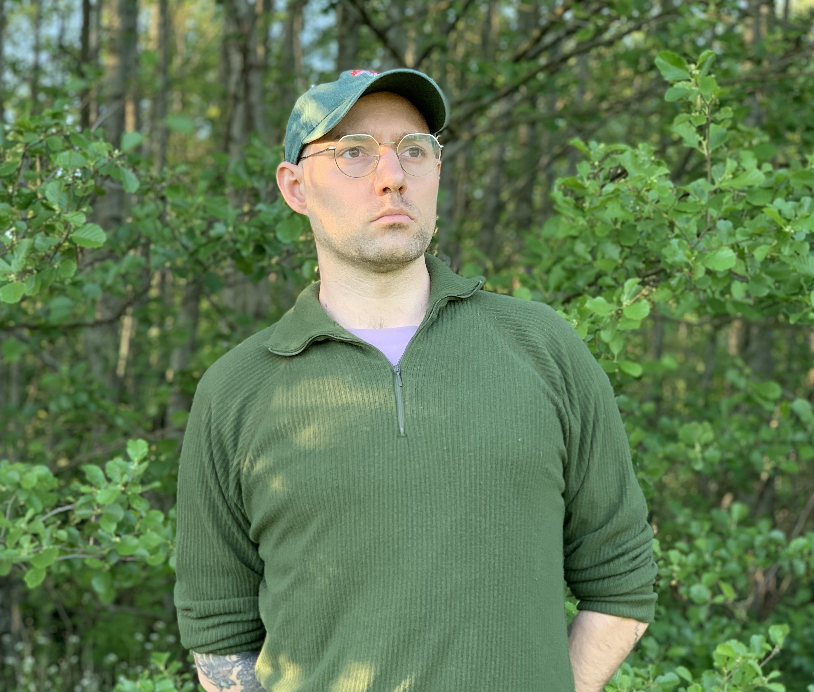 Porträtt på artisten SUNWALD. Han står i en skog framför gröna träd iförd en grön tröja och grön keps.