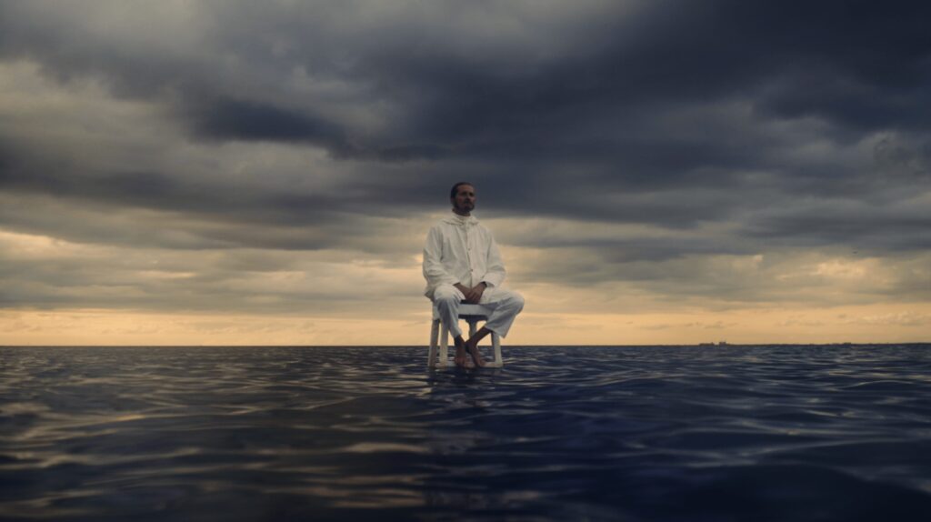 En person i vita kläder som sitter på en vit pall mitt i havet på vattenytan. Runt om personen syns havet och himlen som skiftar mellan ljusa och mörka färger.