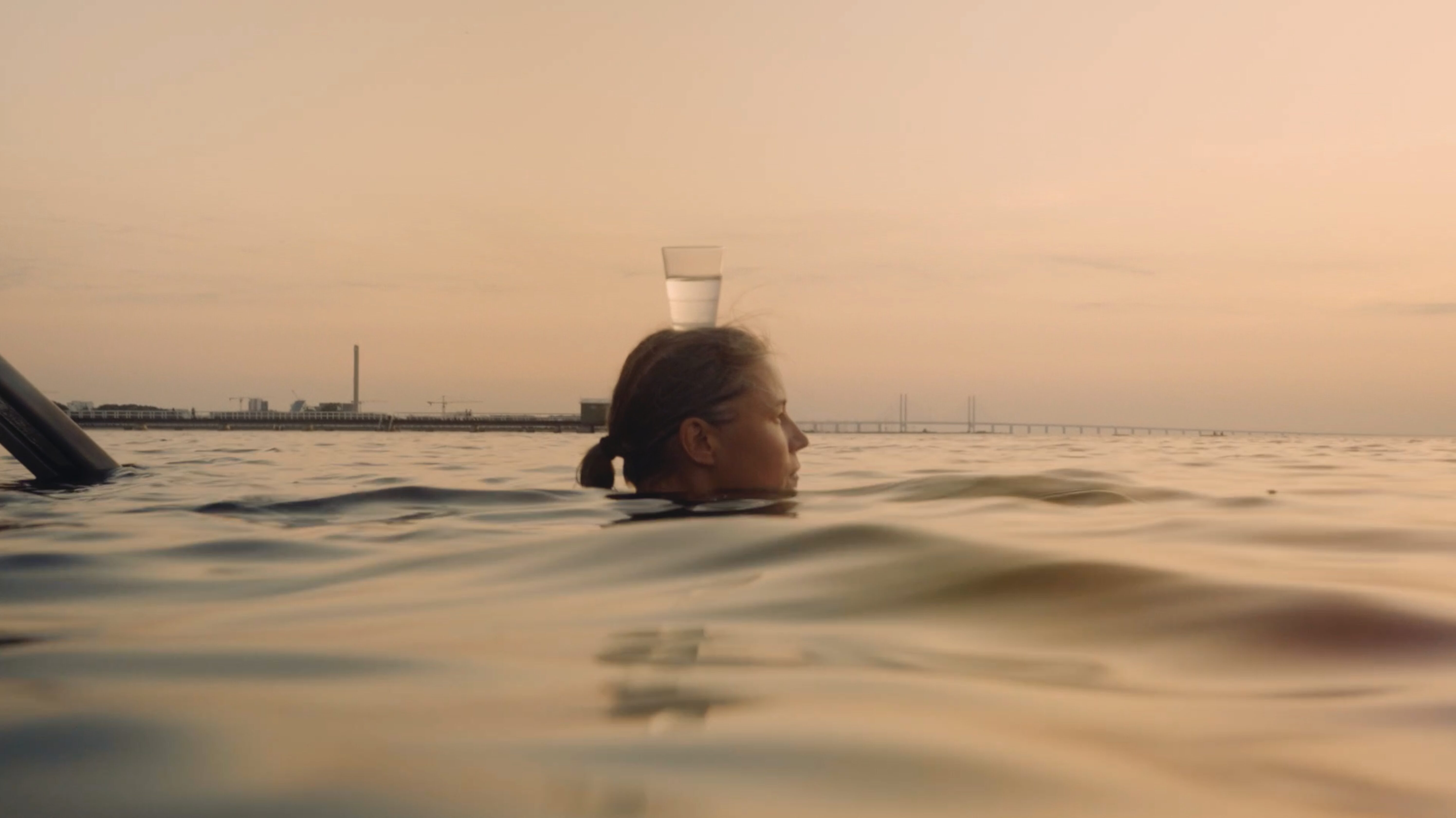 En person som simmar i havet i solnedgång, man ser bara personens huvud ovanför vattenytan. På huvudet balanserar personen ett vattenglas. I bakgrunden av bilden skymtar öresundsbron och några byggnader.