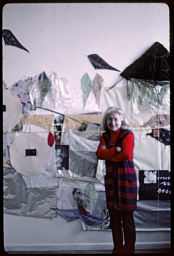 Lite suddigt porträtt av konstnären Lenke Rothman framför en vägg fylld med textila verk.