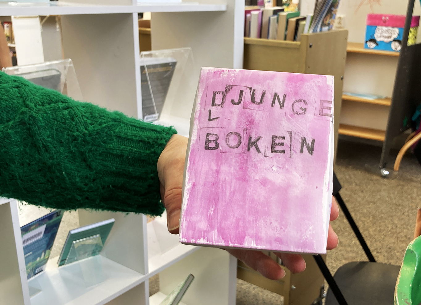 En arm med en grön tröja håller i en rosa bok i handen.