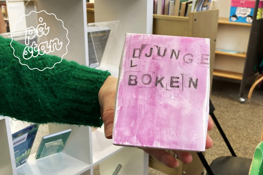 En arm med en grön tröja håller i en rosa bok i handen. En sticker med texten "på stan" i höger hörn.