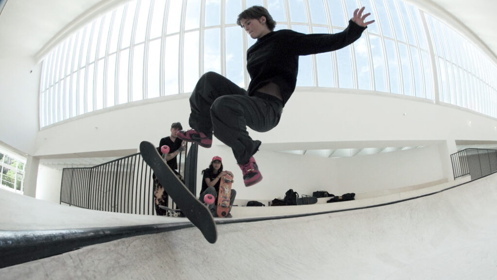 Skateboardåkaren Stella Knutagård gör ett trick i KOO JEONG A's skulptur OooOoO Malmö. Hon är klädd i svarta kläder med röd-svarta skor. Konsthallens stora takfönster syns i bakgrunden.