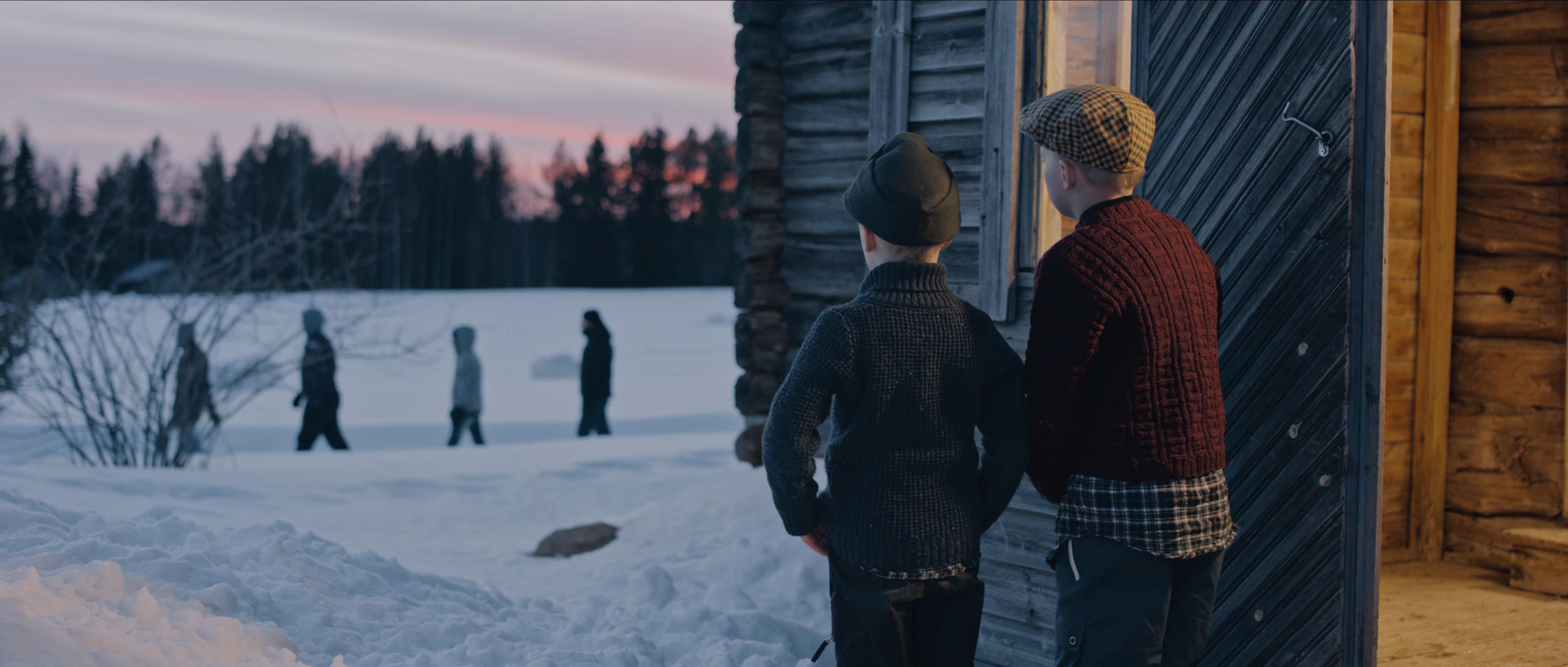 Två pojkar i gammaldags kläder står utanför ett gammalt hus omgivet av snödrivor och tittar ut på några förbipasserande människor.