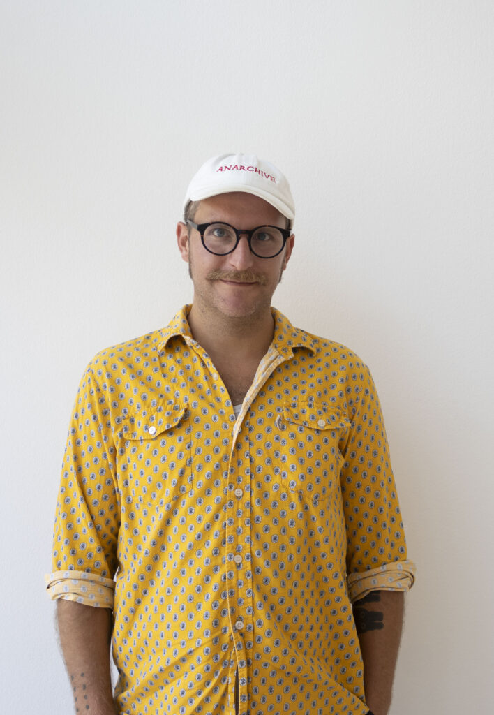 "Portrait of curator Matthew Post. He is wearing a white cap with red text that reads 'Anarchive,' a yellow short-sleeved patterned shirt, and thick black glasses frames."