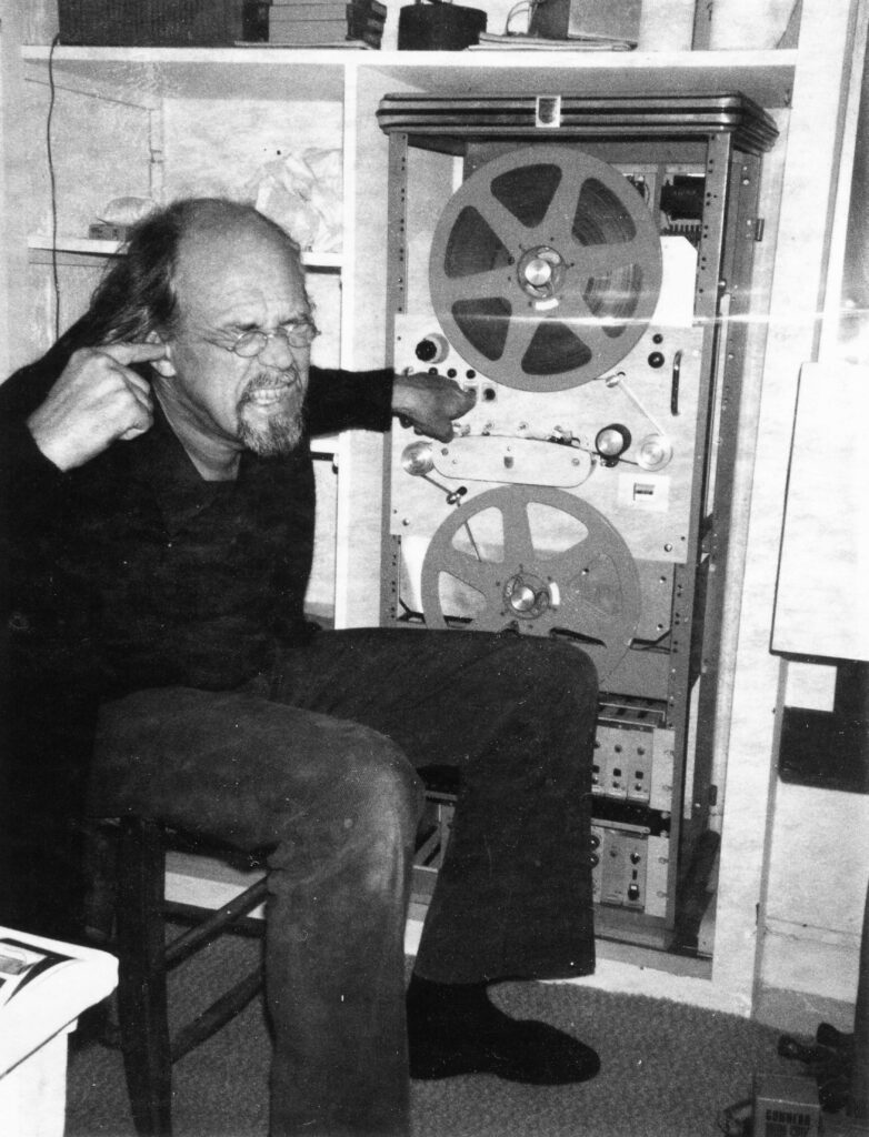 The artist Lars Fredrikson stands in front of a sound-reproducing machine, grimacing and covering his ears. The image is in black and white.