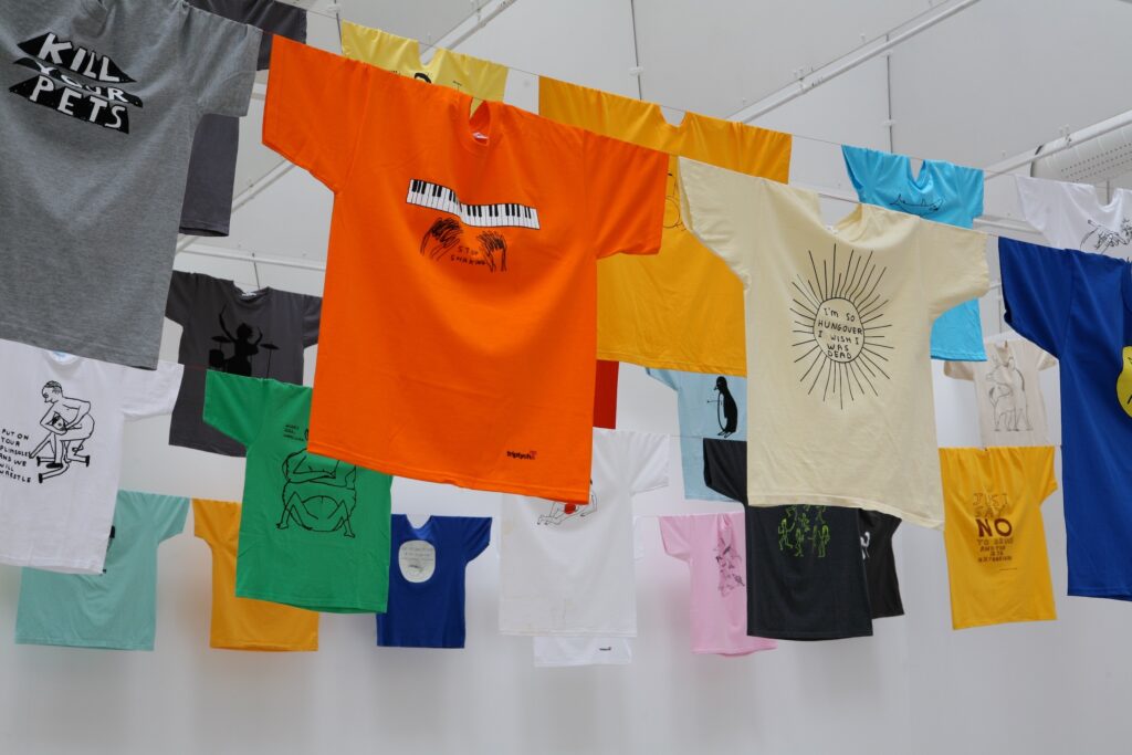 Färgglada t-shirts med tryck hänga i tvättlinor i taket på konsthallen. Tröjorna har tryck som ser handritade ut av konstnären David Shrigley