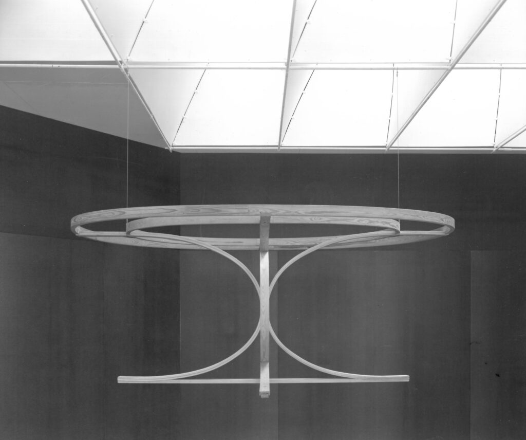 A sculpture in the form of a floating table suspended from the ceiling. The table is constructed of curved wood in rounded shapes but lacks a tabletop at the top.