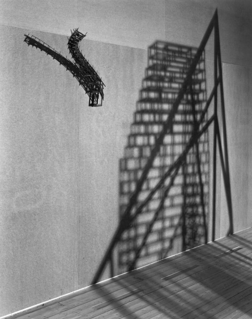 A wall-mounted sculpture extending outward, floating. Beside it, shadows of other sculptures.