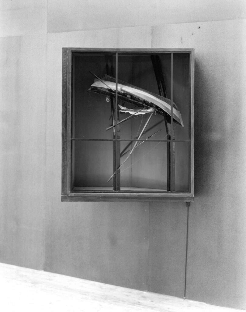 An image of a window frame protruding diagonally from a wall. Tree branches and a model boat emerge from it.