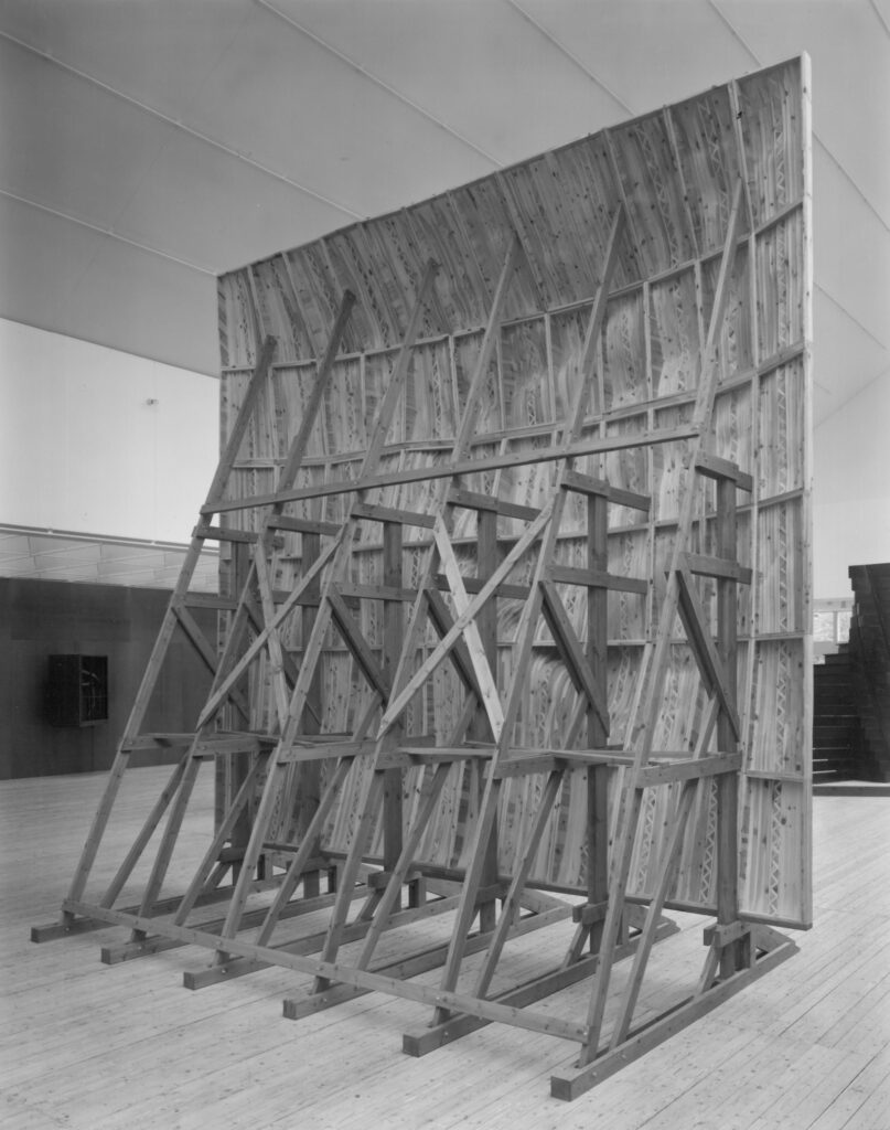 The backside of an undulating wall mounted on a wooden frame standing on the floor. The wall is made of joined wooden pieces and has an uneven surface resembling a wave. The construction consists of massive wooden beams.