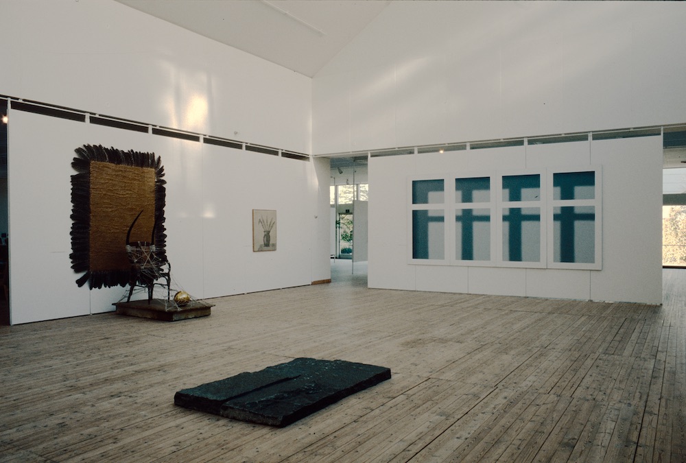 Ett ljust rum i en konsthall med ljusa väggar och trägolv. På väggarna hänger flera konstverk i olika storlek och former. I mitten av rummet ligger en skulptur placerad på golvet.