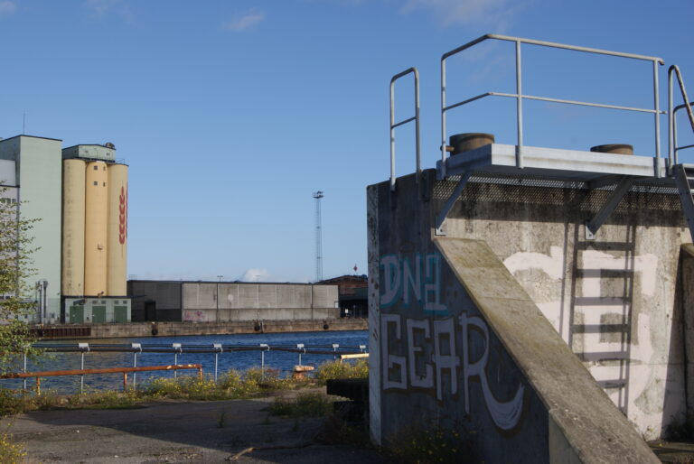 En industriell hamnmiljö med silobyggnader i bakgrunden. I förgrunden syns en betongkonstruktion med graffiti och ett metallräcke ovanpå.