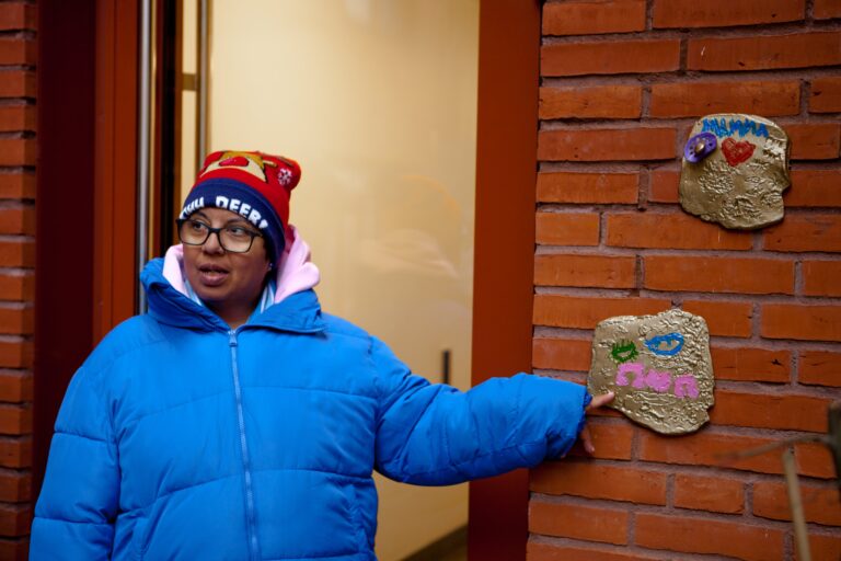 En person iklädd vinterkläder står framför en tegelvägg och pekar på två konstverk som hon gjort, bestående av lerplattor som hon målat på.