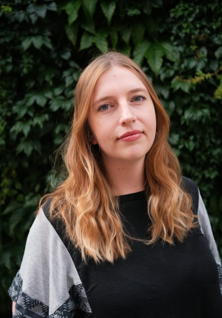 Porträttbild utomhus mot grönskande bakgrund. Lucy har långt rödblont hår, tittar in i kameran med ett litet leende och bär en t-skjorta i svart och grått.
