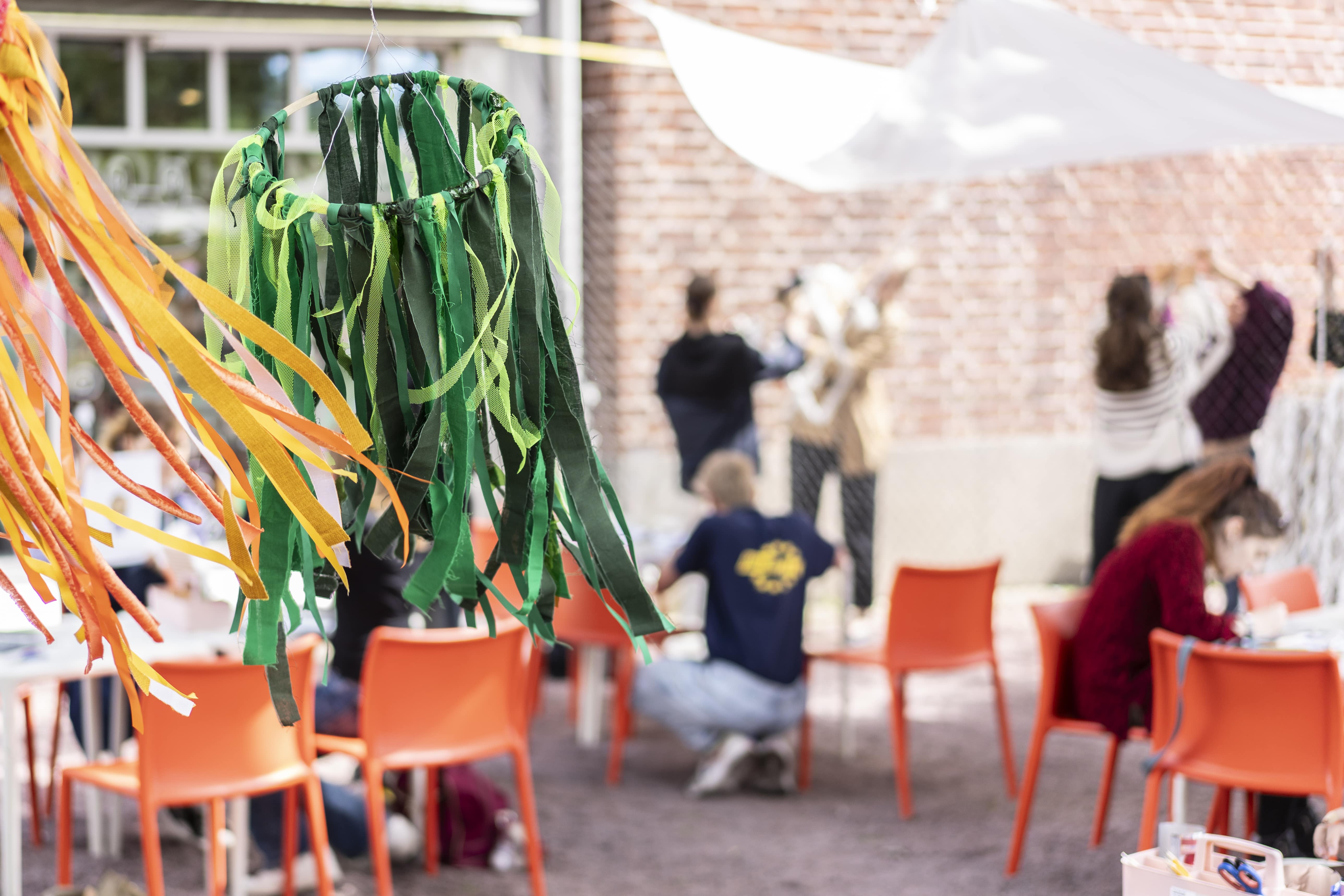Bild tagen på konsthallstorget från en workshop under sommaren. Några ungdomar i bakgrunden målar, i förgrunden färgglada tygdekorationer som fladdrar i vinden.