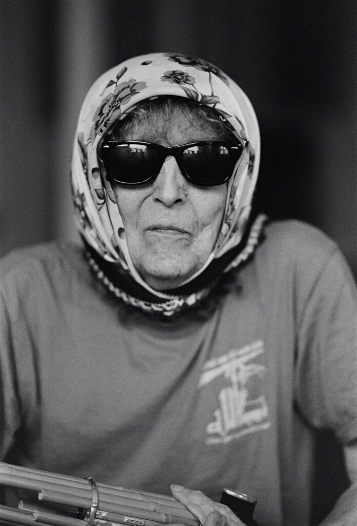 Svartvitt porträtt av CC Hennix i solglasögon och schalett. I nederkant skymtar man ett instrument.