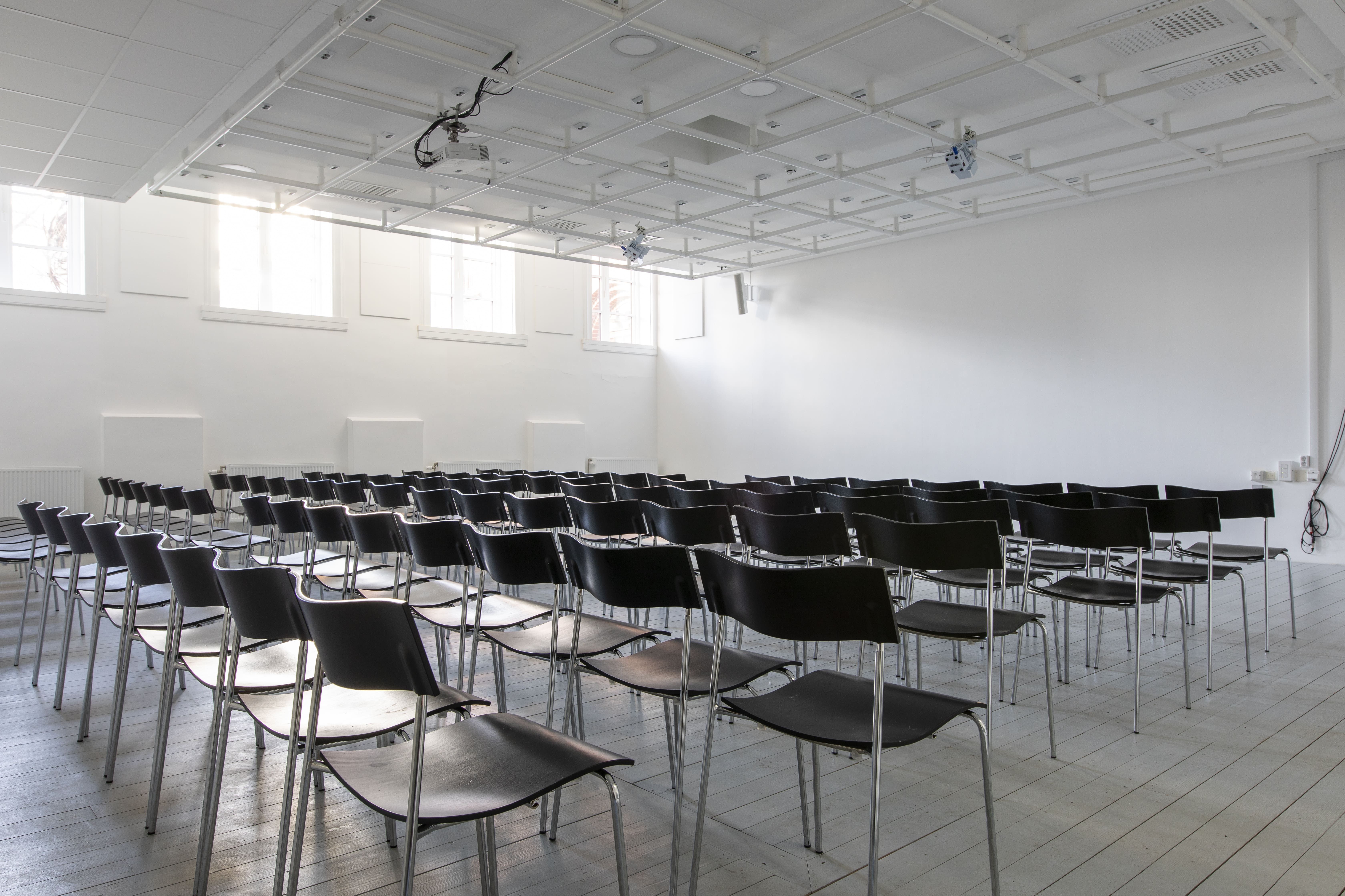 En evenemangslokal med 90 stolar uppradade. Väggarna är vita, golvet av trä och i taket hänger en projektor.