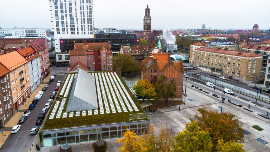 Konstallen fotad från luften så man ser ljussjakten i taket. Bakom konsthallen syns en kyrka och en hög kontorsbyggnad.