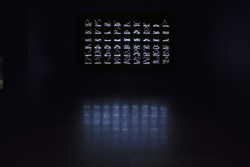 A light installation made up of small steel figures resembling runes, illuminated by LED lights in a dark room.